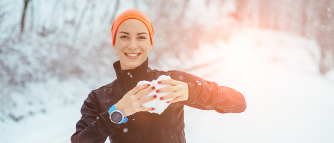 Donna all’aperto in un paesaggio innevato mentre pratica attività fisica, vestita con abbigliamento termico e copricapo. Sorride mentre tiene della neve tra le mani, formando un cuore.