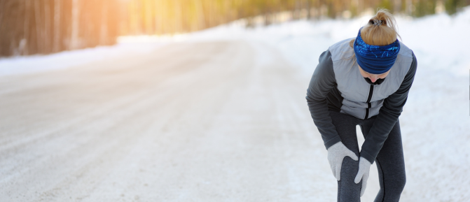 Donna all’aperto su strada innevata che si tiene il ginocchio durante l’attività fisica, immagine rappresentativa di dolori muscolari e articolari accentuati dal freddo.