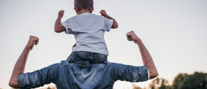 Papà con il figlio sulle spalle al tramonto in un parco, braccia sollevate in gesto di forza e complicità, immagine che rappresenta energia, protezione e dedizione.