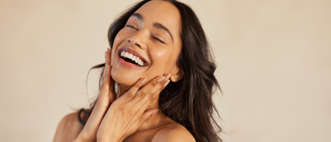 Donna sorridente che si tocca il viso, immagine associata alla cura della pelle dopo l’esposizione al sole e all’importanza dell’idratazione.