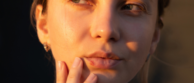 Primo piano di una persona che sfiora il labbro, dove è visibile una piccola lesione compatibile con herpes labiale.