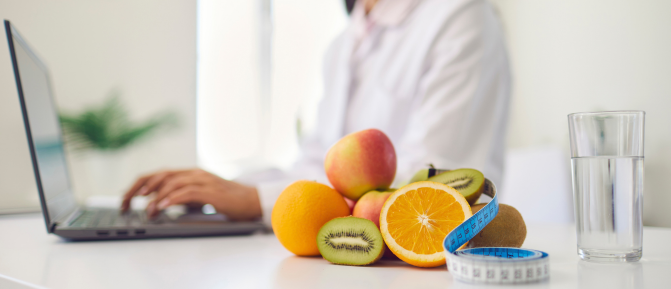 Frutta fresca, metro da sarta e bicchiere d'acqua su un tavolo, con una professionista al computer sullo sfondo, a rappresentare una consulenza alimentare con la nutrizionista in Farmacia.